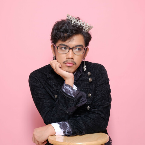 Me in a fashionable goth coat wearing pearl earrings, gem-studded gold tiara and a pair of glasses. My background is rosy pink.