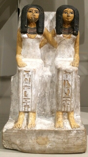 Photo of a statue of two women in Ancient Egyptian white dresses with black, braided hair. They sit side by side, with their arms around each other. 