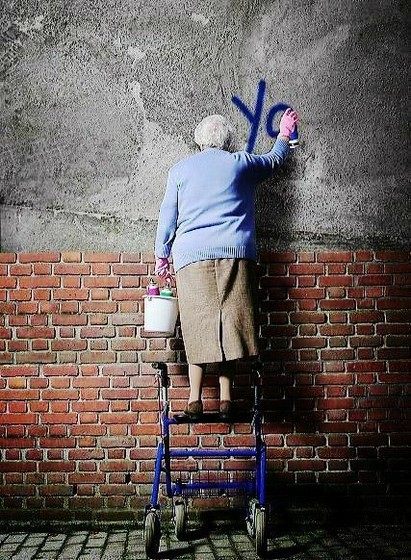 Une grand-mère, debout sur un déambulateur à roulettes, en train de taguer "Yo" sur un mur de béton