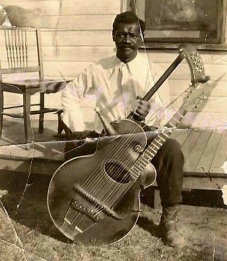 An old B&W photo (in bad condition) of an unknown man sitting on a porch showing his harp-guitar