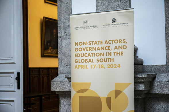 Sign with the title 'Non-state Actors, Governance, and Education in the Global South, April 17-18, 2024. Design on sign is in mustard tones of concentric geometric shapes. In the background is a wood panelled wall with an oil painting of a portrait, and a door ajar.