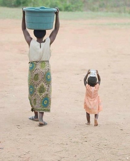 An African woman walks with a large bucket on her head (could be filled with water).
On her right, a little girl holds a mug on her head, like the woman (mother?)