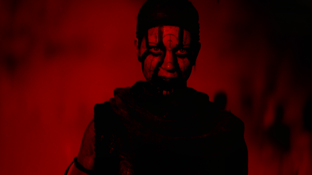 Senua, wearing war paint, looking at the viewer with a red background behind her