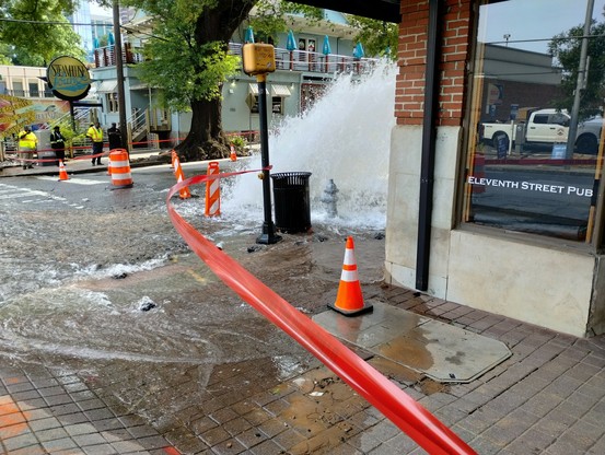 A flooded street corner with water shooting up several feet in the air. The front window of a bar called Eleventh Street Pub is on the corner.