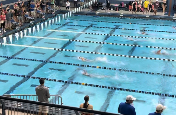 Eight female swimmers are competing in a swim meet. Surrounded by many coaches and spectators.