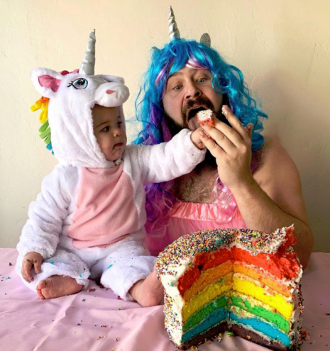 Baby wearing a pink/white unicorn onesie and dad wearing a pink dress, blue wig etc. eating a big multicolor cake