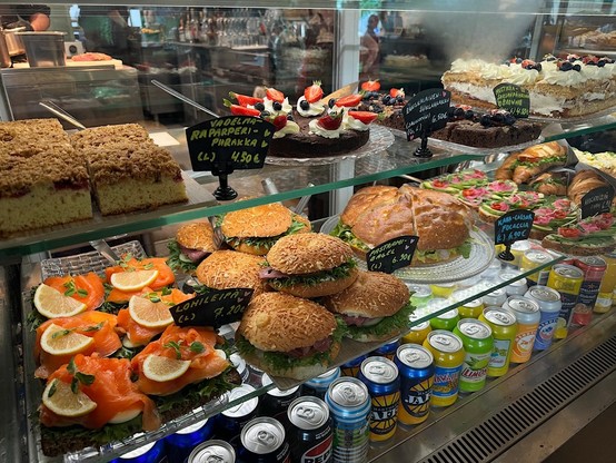 A display case filled with various pastries, cakes, sandwiches, and canned drinks in a cafe or bakery. The items are labeled with prices in euros.