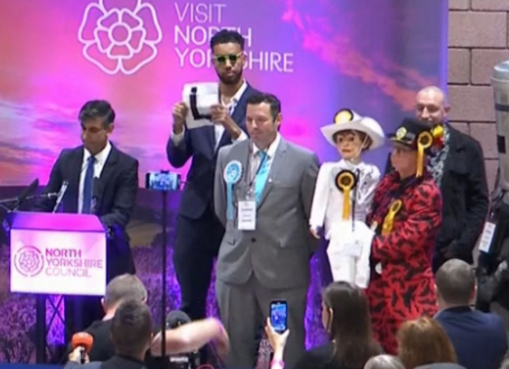 Rishi sunak standing looking defeated at a podium. A person with a funny outfit and a ventriloquist doll stands near him, as does a man with a bin on his head. A guy in sunglasses holds up a sign saying L