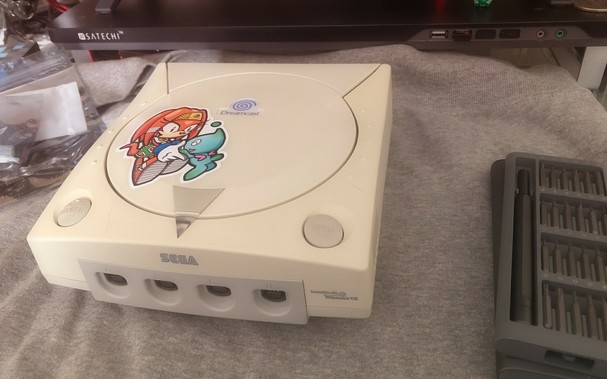 Sega Dreamcast on a desk with a screwdriver set beside it. The Dreamcast has a large sticker on the Disc Tray with Tikal the Echidna and a Chao dancing together. Bagged components of a GDEMU upgrade are beside the Dreamcast.
