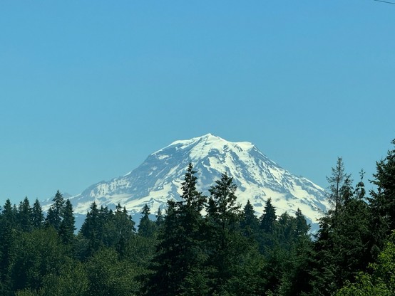 Mount Rainier viewed from State Route 410 in Bonney Lake, Washington, USA. A 14000+ foot stratovolcano, partially covered in snow, rising above evergreen trees. The sky is blue and cloudless. 