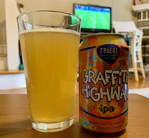 A glass of Hazy IPA and its orange can on a wooden table in a living room. A small TV playing a soccer match is in the background. 