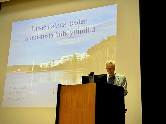 A person stands at a podium presenting in front of a large screen displaying a slide titled 