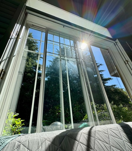 View through a window at an extreme angle looking up from the couch at the sun in blue sky above evergreen trees. Light is streaming through the window, creating rainbow lens effects above the edge of the window. 