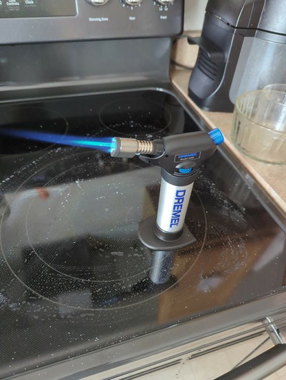 A Dremel torch with flame jutting out, sitting on a glass stovetop.