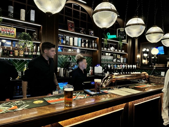 Two bartenders behind a pub bar with drinks and beer taps, illuminated by large pendant lights. Shelves of bottles and a framed sports photo are on the wall behind them.