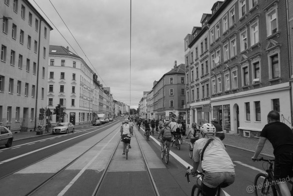 Zwart-wit foto van een groep fietsers, op de rug gezien, die over een lange weg fietsen. In het midden van de weg ligt tramrails. Langs de straat staan huizen van zo'n 4 etages.