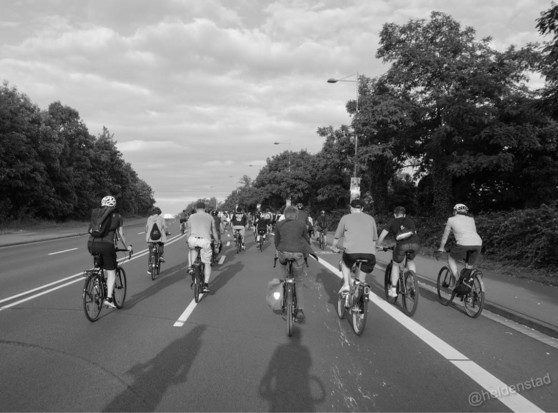 Zwart-wit foto van een groep fietsers, op de rug gezien, die op een brede weg fietsen. De weg loopt iets omhoog naar een brug. Bomen staan aan beide zijden van de weg. Het is half bewolkt maar de schaduwen van de fietser zijn duidelijk te zien.