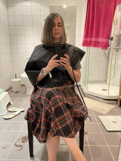 Woman sitting in a chair after a fresh haircut, taking a selfie. Various bathroom objects are visible in the background. A fuchsia towel on a sheet cabin, flip-flops, hair cuttings. The floor is light brown and the walls are white. Woman is dressed in dark grey tshirt and dark tartan skirt. The tartan colors are black, red and some white. Woman has brown hair with grey strands. A black cover is used to catch the cut hair. Woman has a white sports watch.