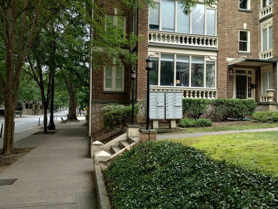 A brick apartment building next to a tree lined street. Very poorly photographed in one window are the two flags.