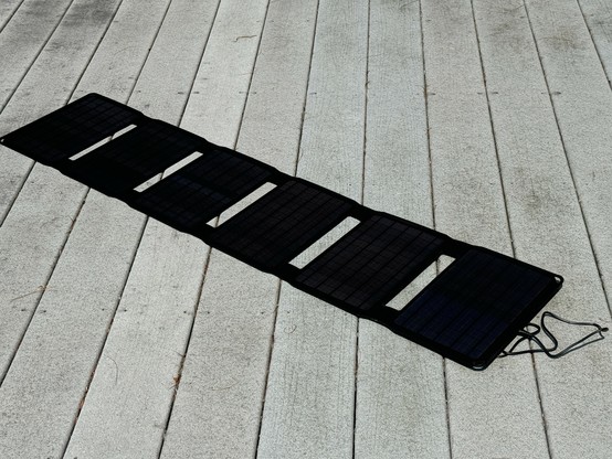 A six-segment portable solar panel charger. It’s black and sits on a grey porch. There is a cable visible on the right. 