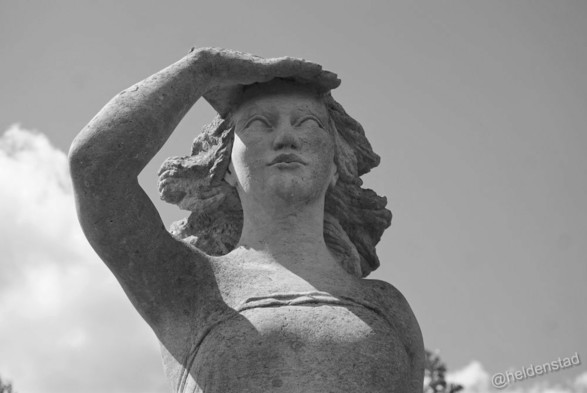 Zwart-wit foto. Close-up van het bovenste deel een standbeeld. Het is een vrouw die haar rechterhand boven de ogen heeft en in de verte tuurt.