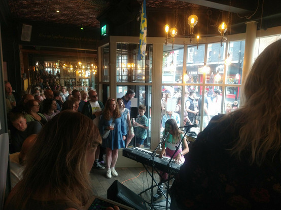 A young woman sings while playing a keyboard, surrounded by an audience in a busy pub. The street outside is also busy, seen through the large window behind her.