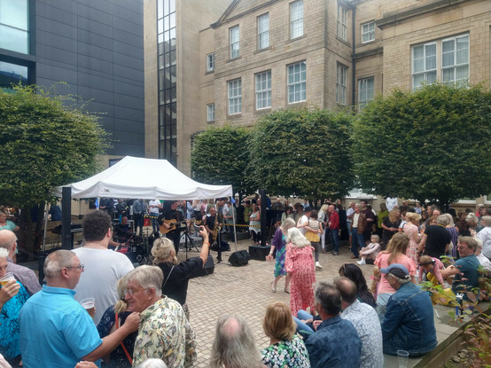 A band under a canopy in a city centre plaza, with a keyboard player, drummer, guitarist and saxophonist. They are surrounded by lots of people, except for an empty area in front of the band where a few people are dancing.