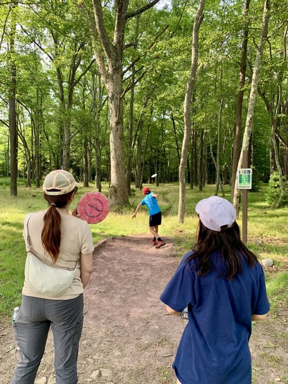 A boy wearing a blue t-shirt and red hat is throwing a disc into the forest. His sister and mom are waiting for their turn.