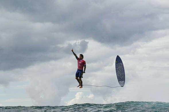 Photo of Brazil's Gabriel Medina rising above the ocean surrounded by white clouds; right arm raised, finger pointing skyward his surfboard tethered to his ankle rising  vertically in the air beside him. Photographed by Jerome Brouillet- AFP Getty Images in Teahupo'o,  Tahiti.