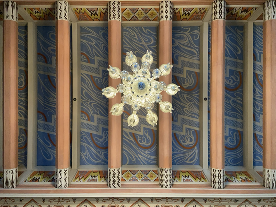 View of a chandelier from underneath with yellow globes poking out from around a central base. The ceiling has brown beams and a patterned blue background. 