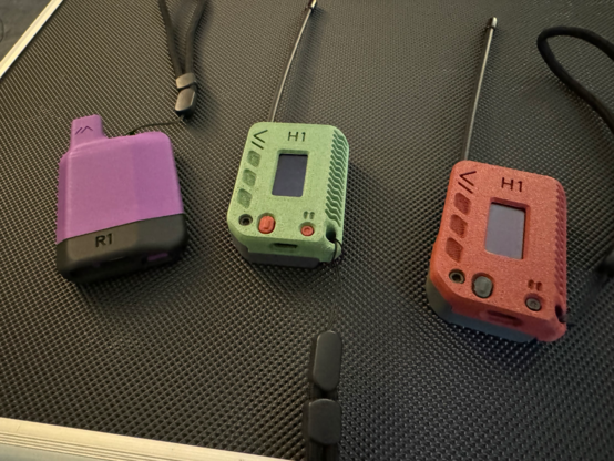 Three LoRa Meshtastic devices. One is green, one is purple and one is red. The red and the green ones have little screens and 6” antennae. The purple one looks like a vape device.