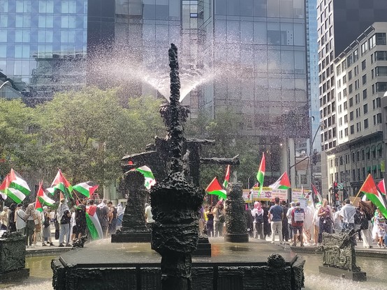 A black water fountain in front center, many people with big Palestine flags in front of it, business buildings and trees in background 