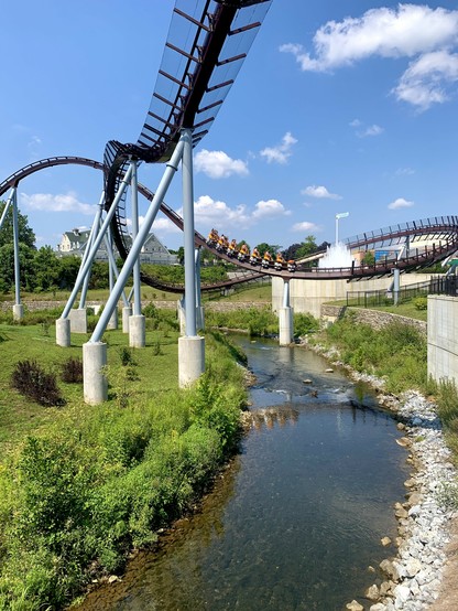 Chocolate colored roller coaster track over a small creek. The blue sky in the background, the creek runs through grassy area of Hershey Park. A seven-compartment roller coaster is running over the creek.