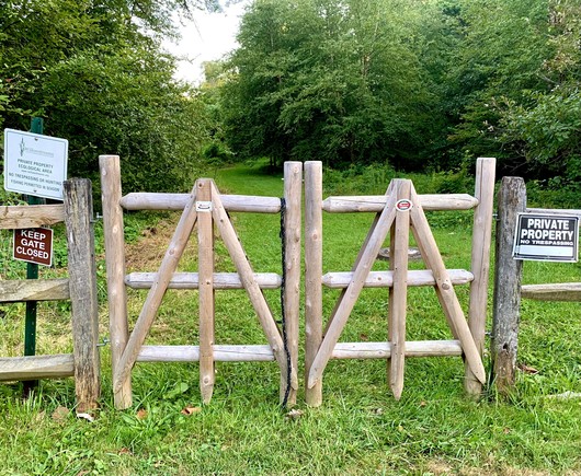 A wooden gate leads to a grassy trail surrounded by lush, vivid green woods. There are three signs: one reads 'Private Property,' another says 'Keep Gate Closed,' and the last states 'Fishing Permitted.