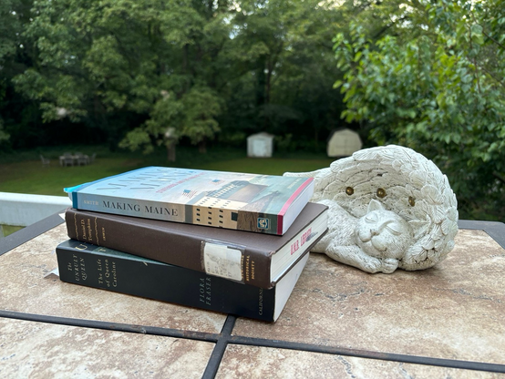 A stack of three books on an outdoor table with a lawn and trees in the background