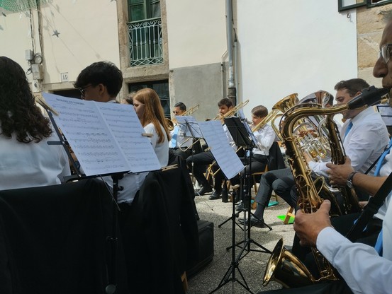 Picture of a music band playng in the street. They are seated in chairs, playing wind instruments (saxophones, trombones, etc), in front of sheet music, wearing black trousers and white shirt. Looks like a popular Galician party.