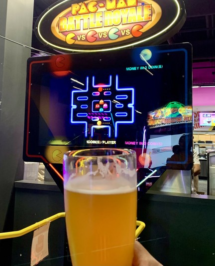 Holding a glass of IPA in front of the Pac-Man video game in an arcade.