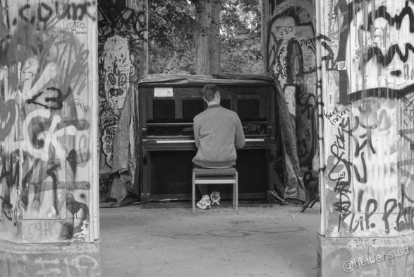 Zwart-wit foto van een persoon die piano speelt in een betonnen paviljoen. Door de compositie zijn links en rechts naast de muzikant enkel de pilaren te zien. Deze staan vol met graffiti.