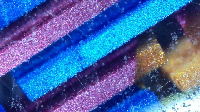 Photo of a fountain pen under a microscope.

There are rectangular bars of color with a square cross-section going from the lower left to upper right. They are mostly blue and purple, with one partially gold. They appear to be suspended in clear resin.

Some surface scratches and pits are visible.