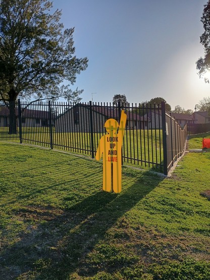 A yellow sign in the shape of a person pointing upwards, with the words plastered over its body saying: Look up and live! In the background are some buildings with a bright blue sky and the sun above.