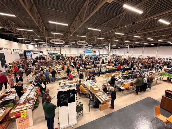 A large indoor event featuring numerous tables filled with books and records. Many attendees browse the aisles and engage in conversations. Display signs indicate various sections, and there are several booths in the background. The atmosphere is busy and lively.