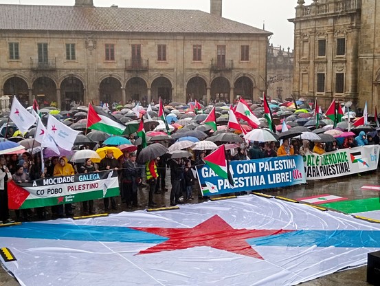 Foto de manifestación na praçaa da Quintana. Praça entre prédios antigos de pedra. Centos de paraugas, muitas bandeiras de Palestina e do Líbano e de Galiza Nova. Faixas de Galiza Nova (