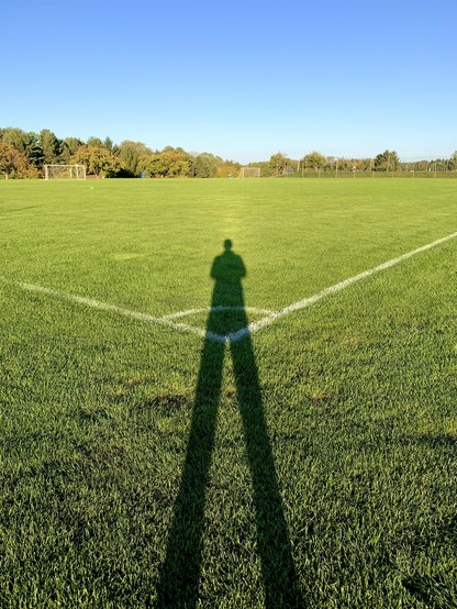 A person is taking a photo of their shadow cast on the bright green grass of a soccer field. Beyond the field, a line of woods stretches under a clear blue sky.