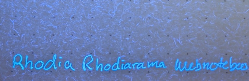 Photo of written text on paper. The paper has a 5mm dot grid. The surface of the paper has a purple cast. There are many bright purple glowing fibers visible throughout the paper surface.

The text is glowing very bright blue and reads 