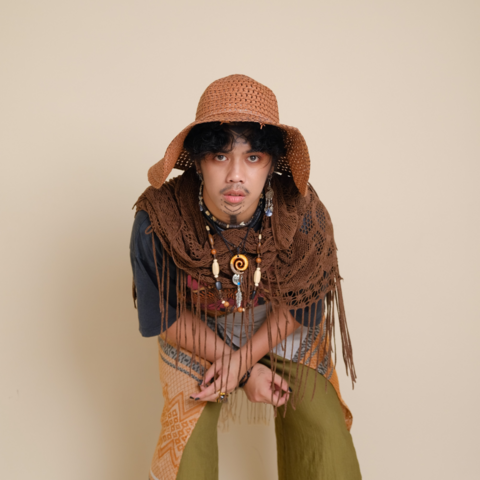 Me in my Boho outfit. My chin is painted with black downward chevron markings. My neck has a thick black line going down through it. I am wearing a variety of wooden and metal necklaces, while also draped with a netted scarf. My head is covered with an earthen orange sunhat. I am wearing a flowy pair of moss green pants overlaid with a makeshift skirt made out of patterned pashmina scarf.