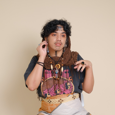 Me in my Boho outfit. My chin is painted with black downward chevron markings. My forehead is painted with black flourishes and a triangle. My neck has a thick black line going down through it. I am wearing a variety of wooden and metal necklaces on top of my black print shirt, while also draped with a netted scarf. I am wearing a makeshift skirt made out of patterned pashmina scarf held together by a woven yarn belt.
