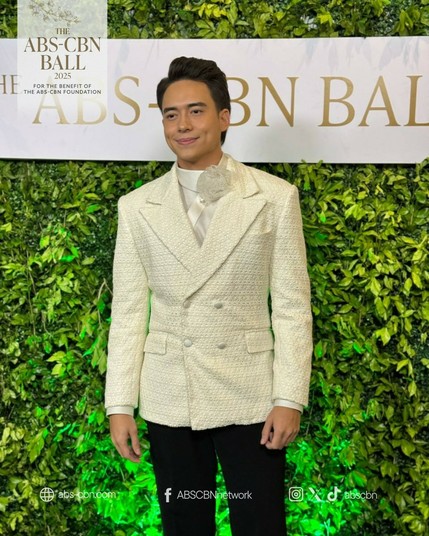 Jameson Blake in a white double-breasted suit on top of a satin white shirt accentuated with a tulle puff where a bow should be and is placed to the right side of his neck. He wears black pants.