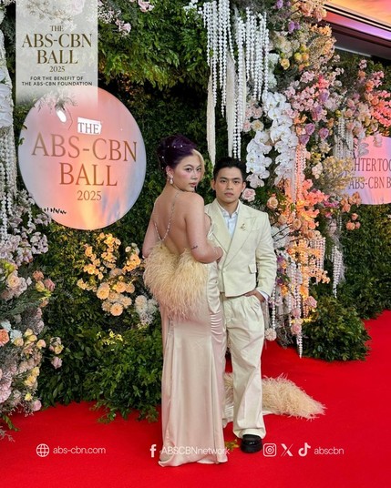 Carlos Yulo wearing an all-cream suit and pants. Chloe San Jose wearing a backless long gown decorated with fur at her back. Her hair is dark burgundy, with her bangs colored almost platinum blonde.