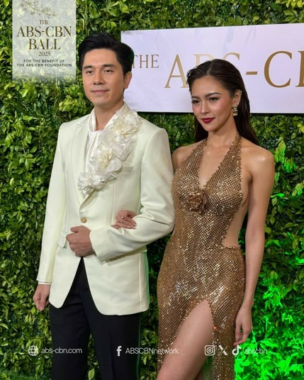 Paulo Avelino in a white tux, with its right collar covered with white flowers. Kim Chiu in a long fluid dress made of bronze-colored metallic material. Her sides and her back are exposed, further emphasizing her figure.