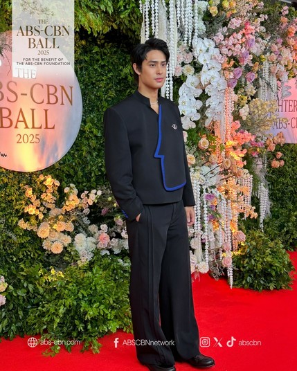 Donny Pangilinan in a modern black suit with wide-leg pants and an asymmetrical blue-outlined flap stands confidently on the red carpet at the ABS-CBN Ball 2025, surrounded by lush floral decor.